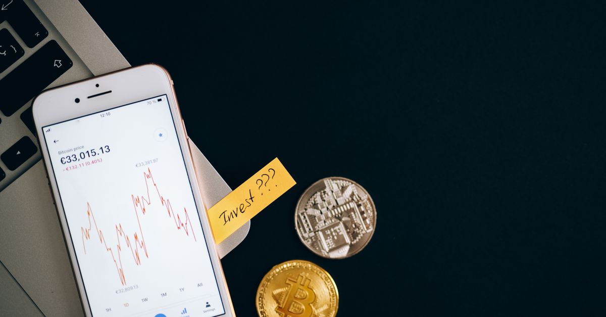 A smartphone displaying cryptocurrency prices beside coins and a laptop, symbolizing investment.