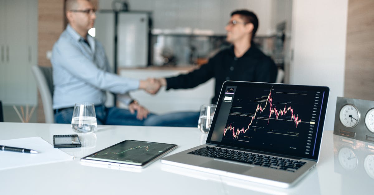 Two businessmen shake hands at a desk with stock market graphs on a laptop.