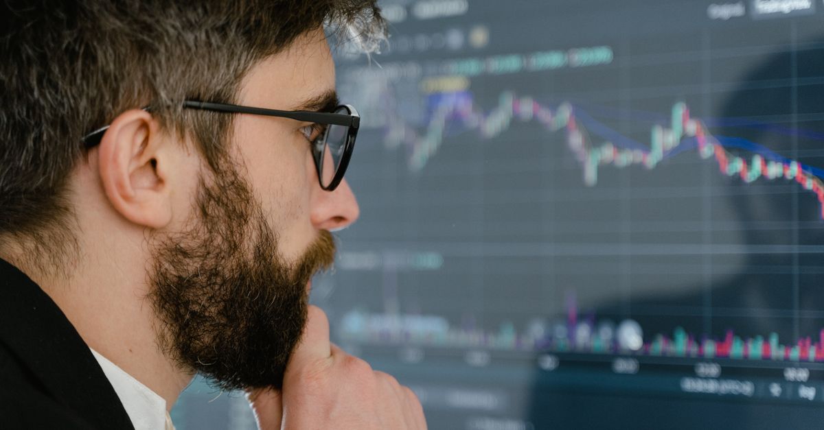 A focused man with glasses studies stock market graphs on a screen, pondering insights.