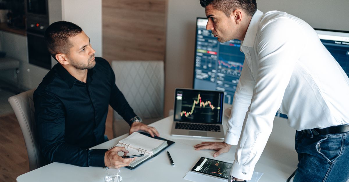 Two businessmen discuss stock market data on screens in a modern office.