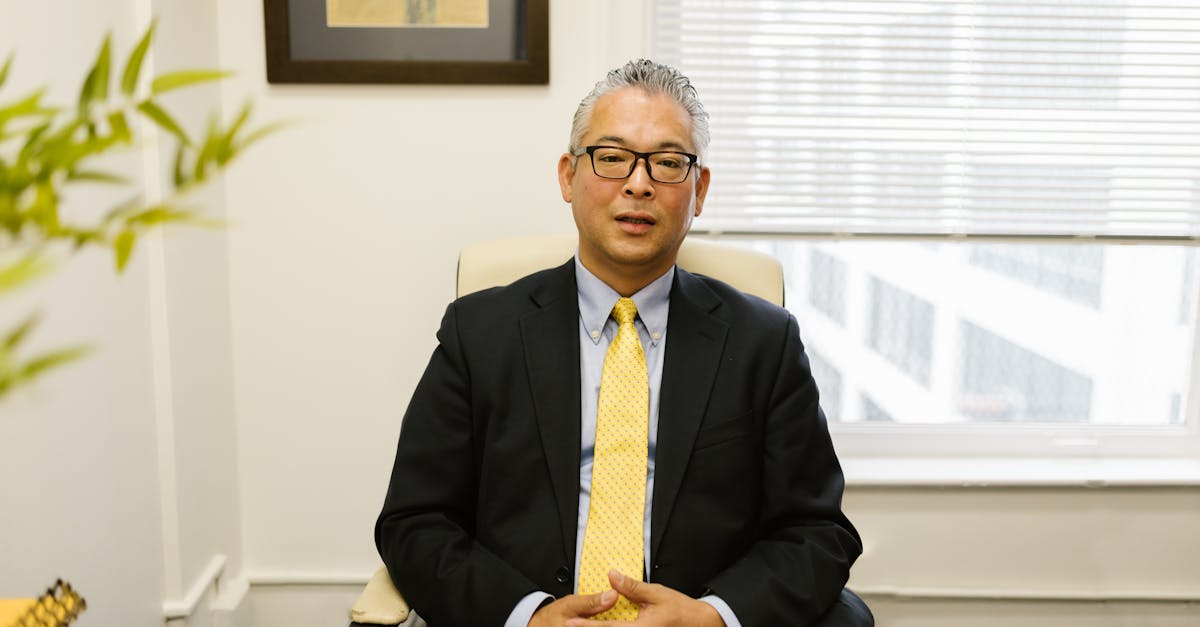 Middle-aged man in business attire sitting at an office desk with a blurred background.
