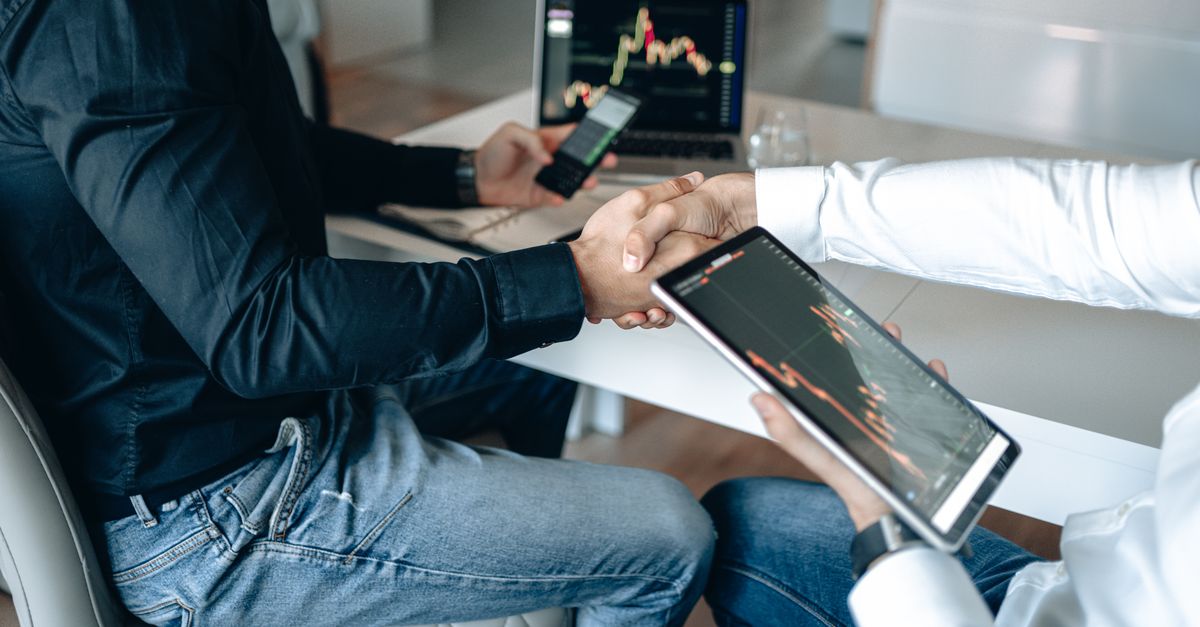 Two business professionals shaking hands with financial graphs on digital devices.