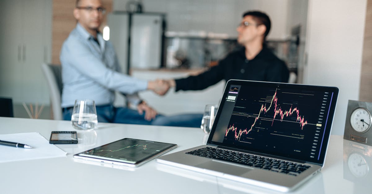 Two businessmen shaking hands in an office setting, with laptops displaying financial graphs.