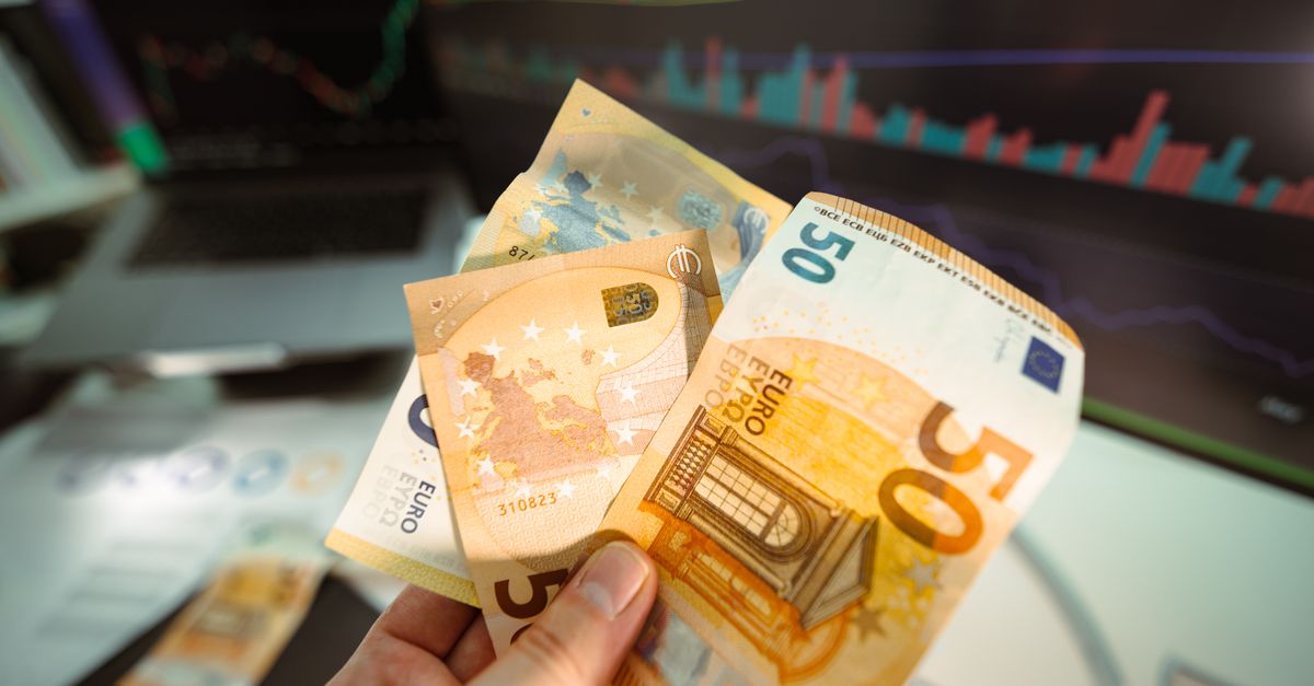 Hand holding Euro banknotes in front of a trading screen showing financial graphs.