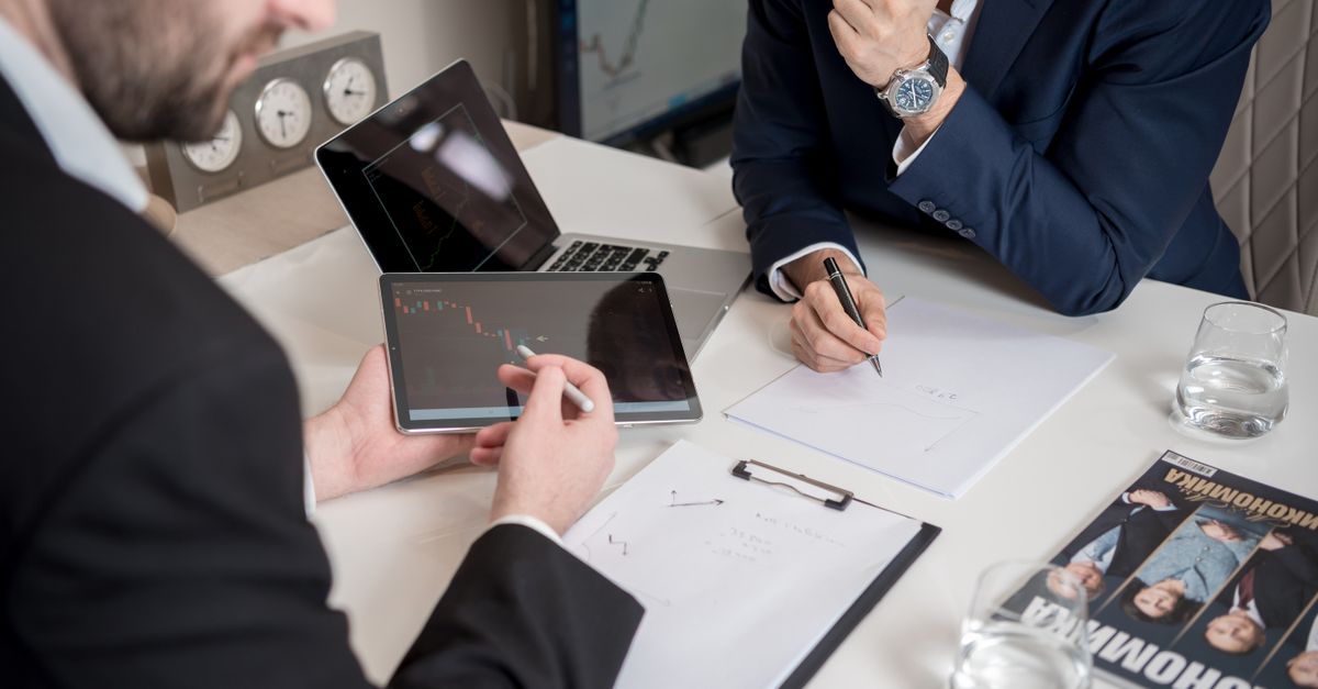 Two businessmen discussing financial charts on a tablet in a modern office setting.