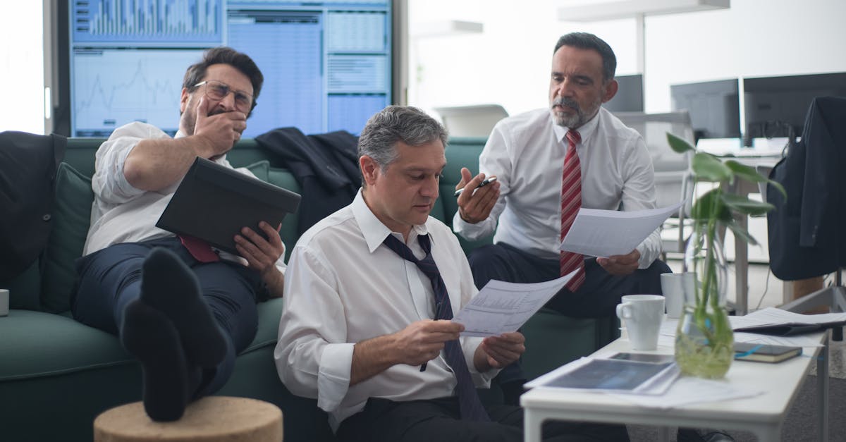 Three businessmen review documents while sitting in an office environment with screens displaying data.
