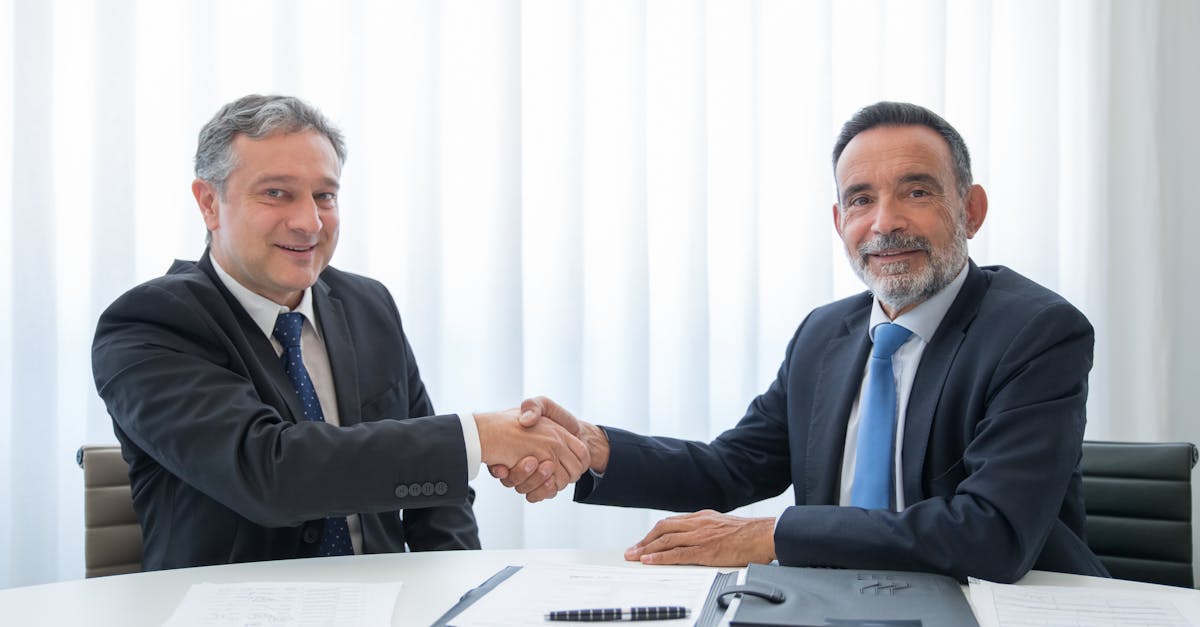 Two businessmen in suits shaking hands at an office table with documents.