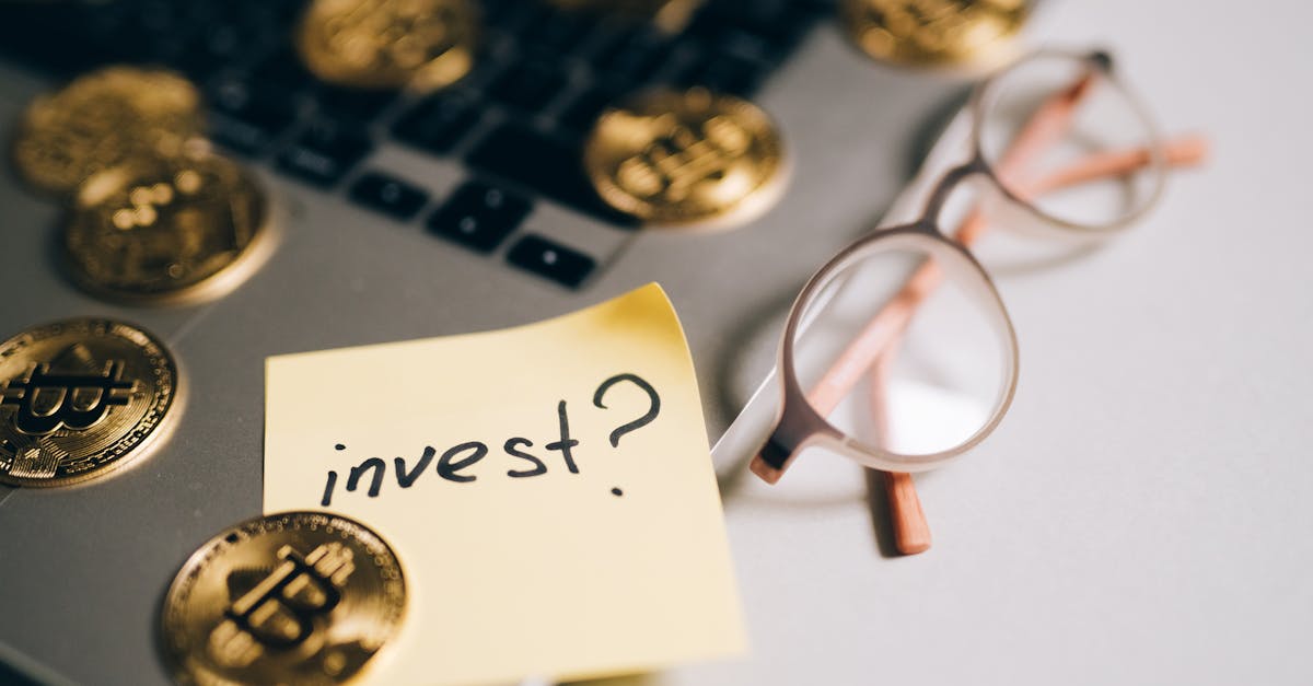 A close-up of bitcoin coins on a laptop with eyeglasses and a sticky note reading 'invest?'