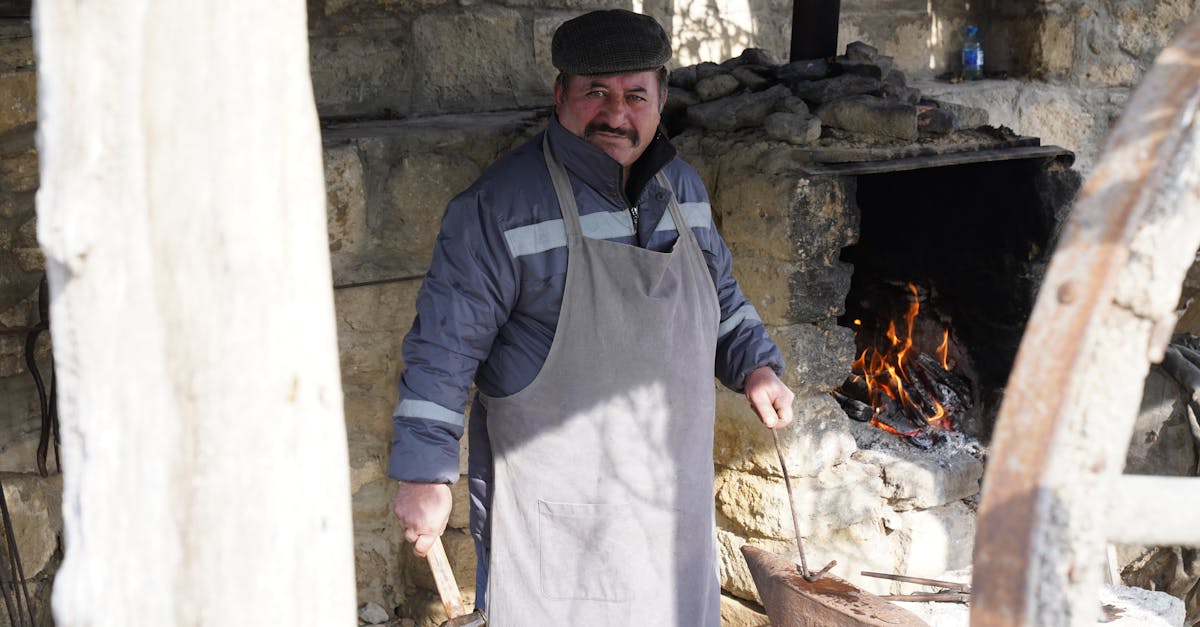 A blacksmith works at a traditional forge outdoors, wearing an apron and holding tools.