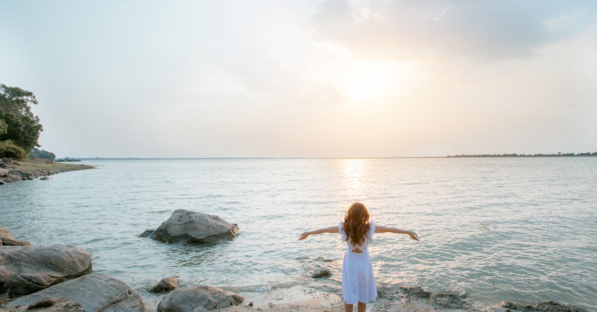 A woman in a white dress stands on a rocky beach, embracing the serene seascape at sunset.