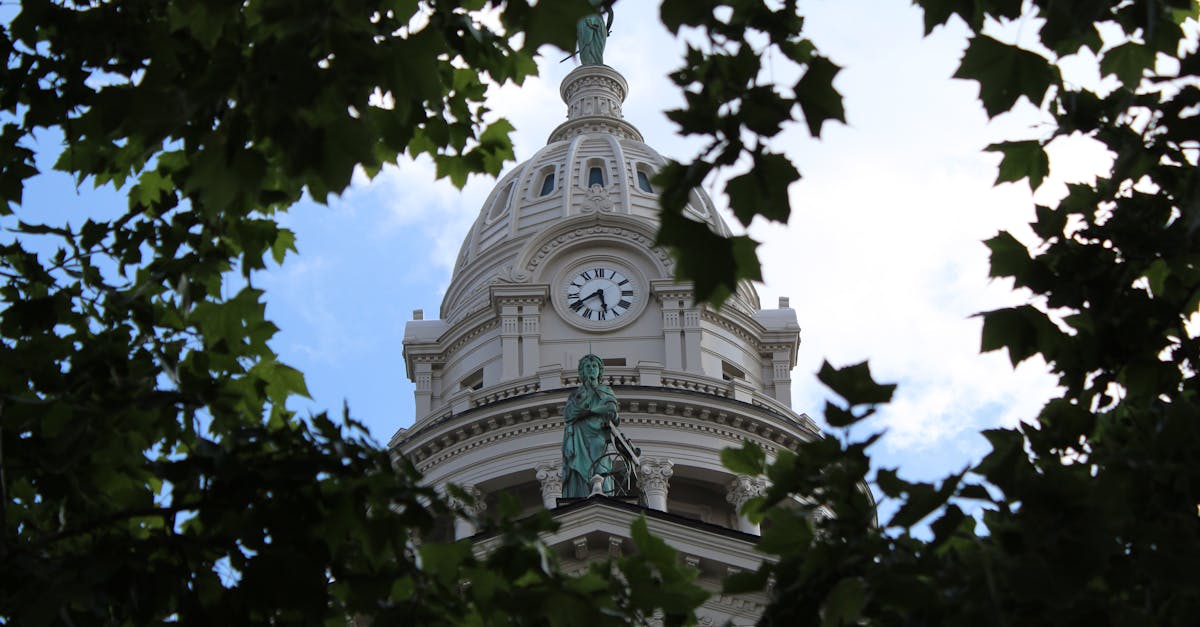 A historic clock tower with a dome and statue surrounded by green leaves against a blue sky in Troy, Ohio.