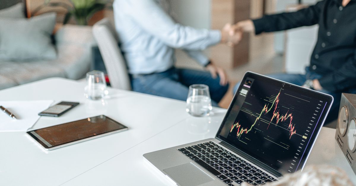 Two professionals shaking hands in an office with data analysis on a laptop screen.