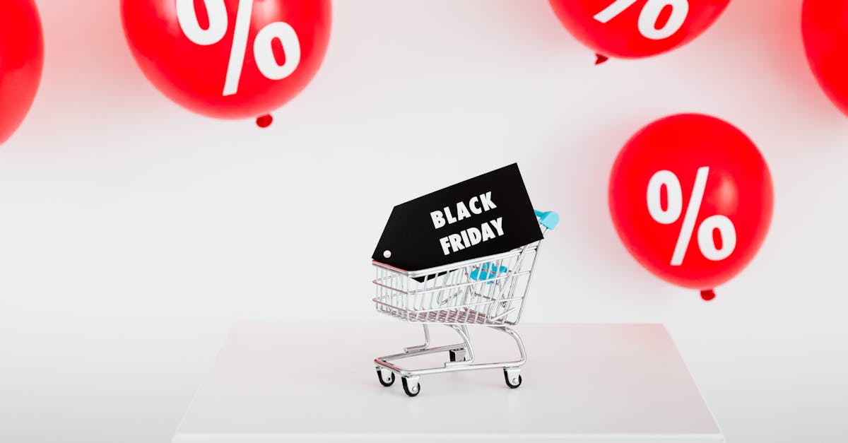 Mini shopping cart with Black Friday tag surrounded by red percent balloons on white platform.
