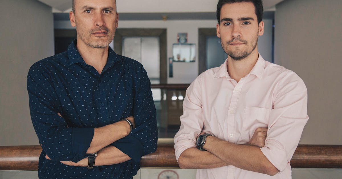 Serious handsome men in casual clothes standing together with crossed arms near glass fence in light corridor while looking at camera