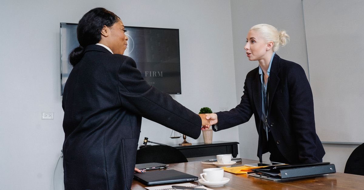 Smiling multiracial women in formal wear standing near table with documents and shaking hands after successful deal