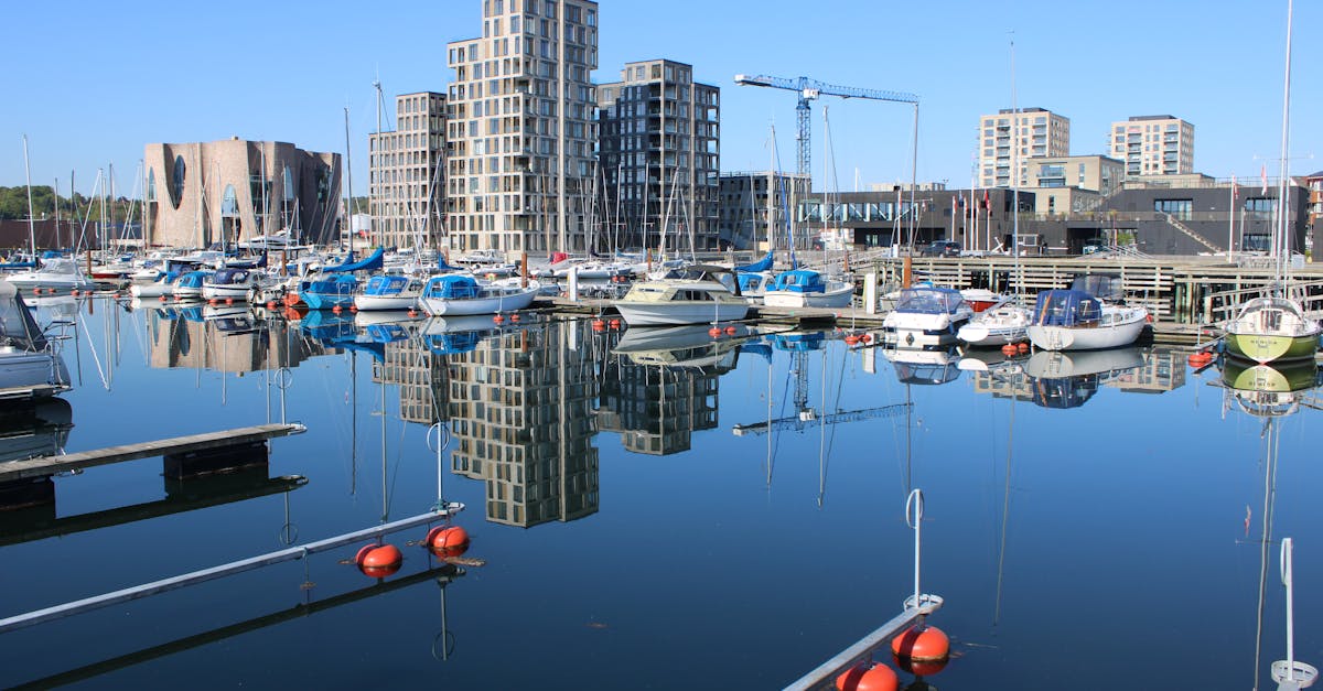 Boats moored in Vejle's harbor, reflecting modern buildings under a clear sky.