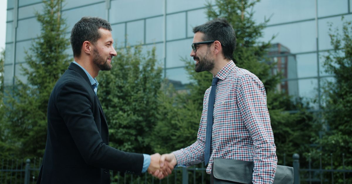 Two men shaking hands outside a modern office building, symbolizing business partnership.