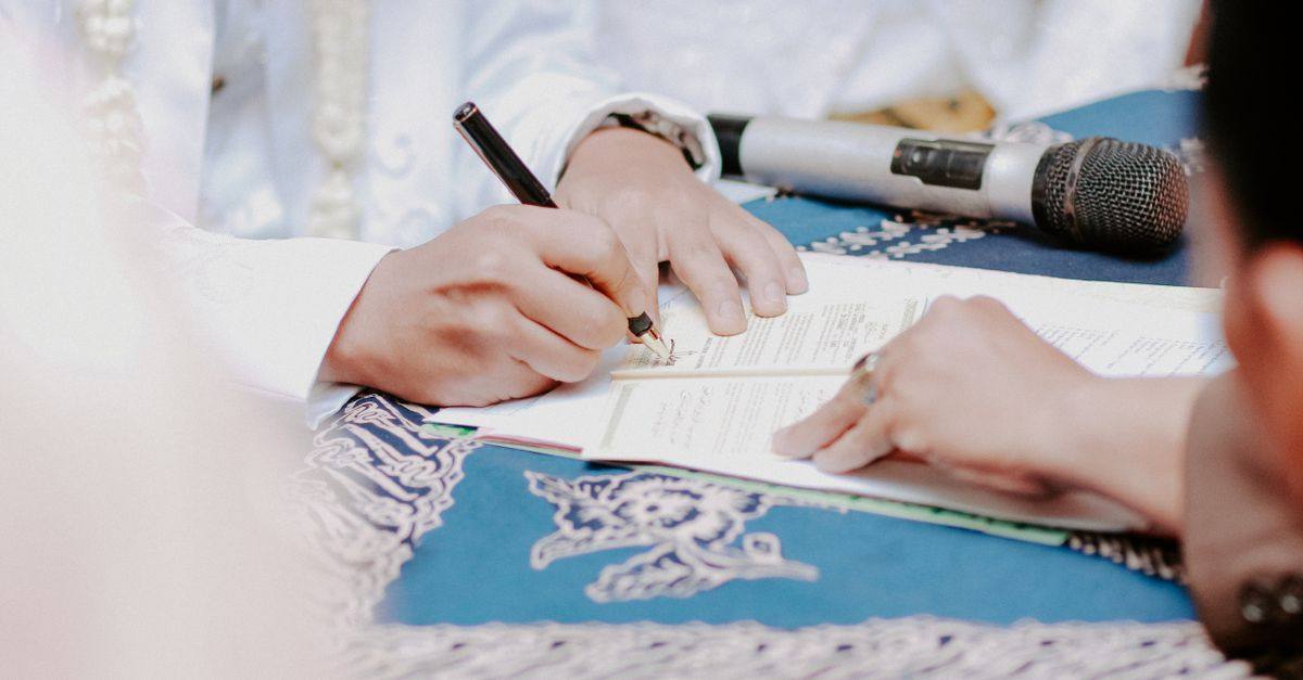 Close-up of hands signing wedding documents with microphone on table.