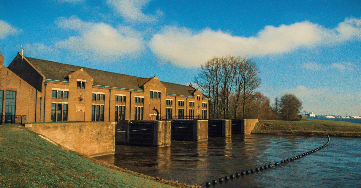A historic pumping station by the river under a clear blue sky, framed by trees.