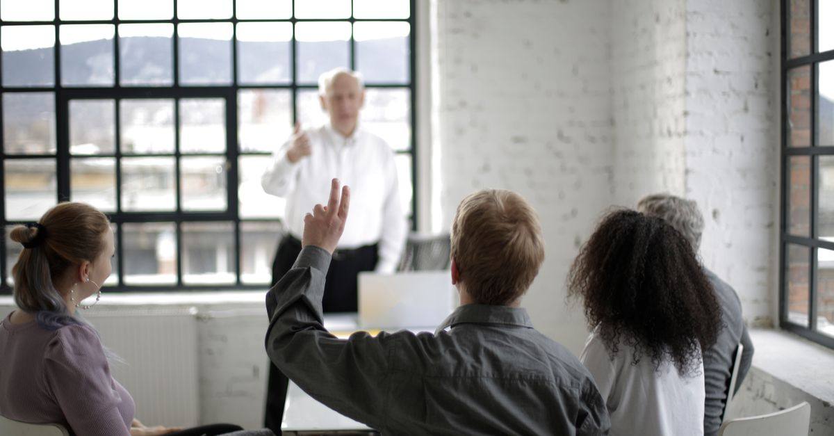 Group of adults in a discussion, with one person raising a hand in a bright office space.