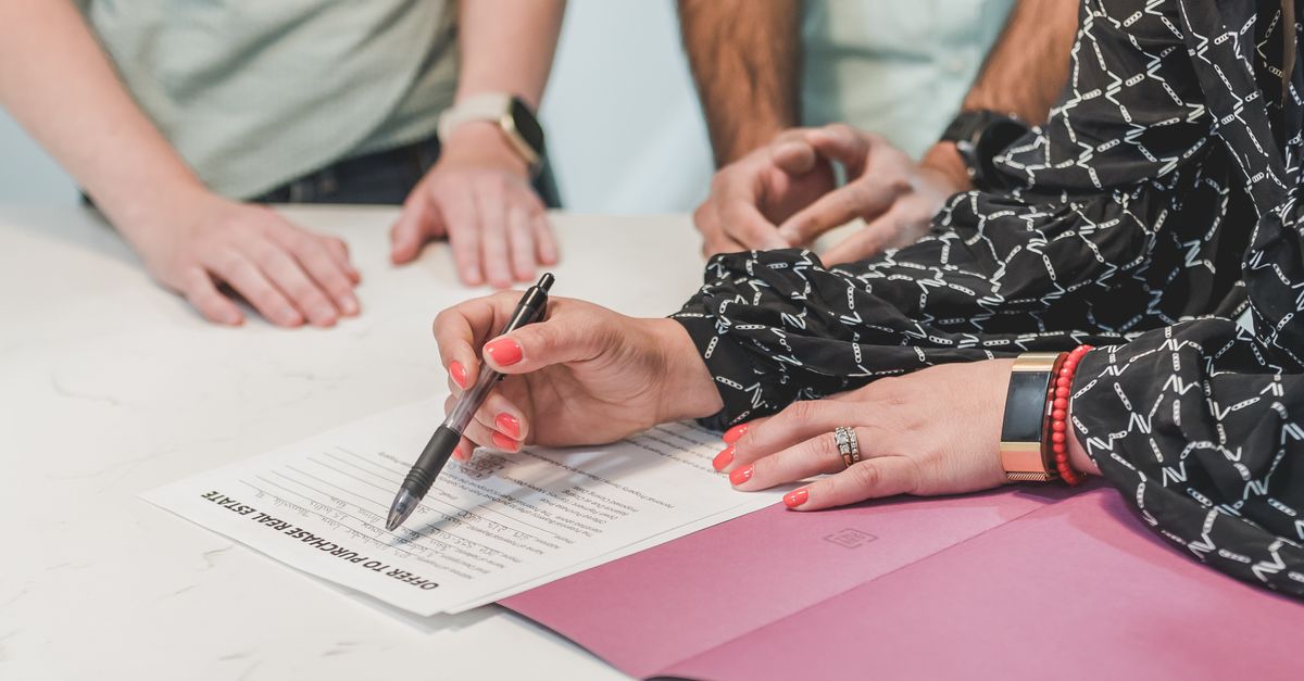 Close-up of hands signing a real estate document at a meeting table with three people.
