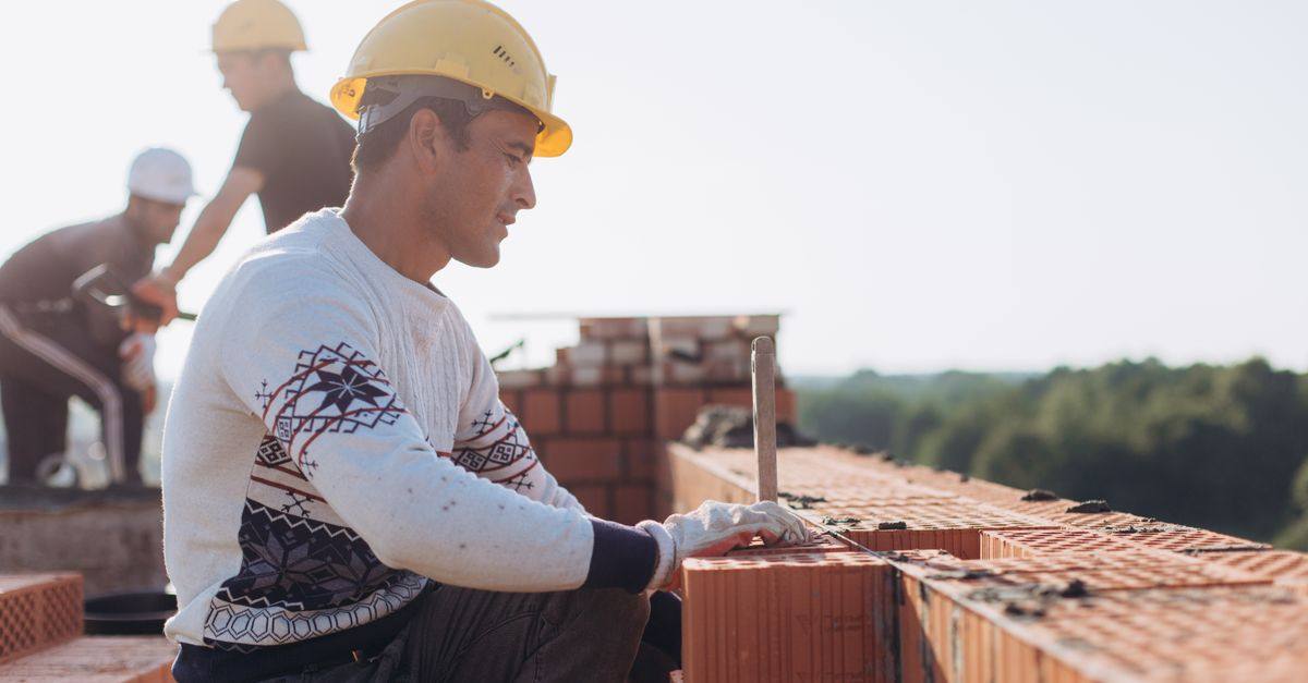 Construction worker laying bricks outdoors in sunny weather with lush greenery in background.