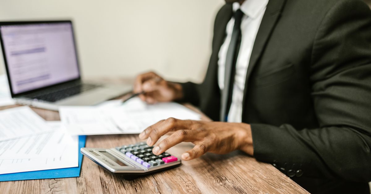 A businessman in a black suit using a calculator at a desk with financial documents and a laptop.