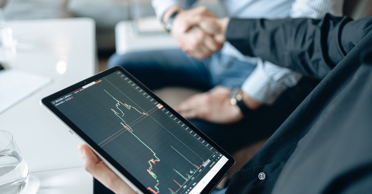 Two adults shaking hands with a stock market chart on a tablet, symbolizing a business deal.