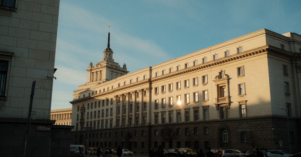 A warm sunset illuminates the National Assembly building in Sofia, Bulgaria, with a clear blue sky and passing pedestrians.