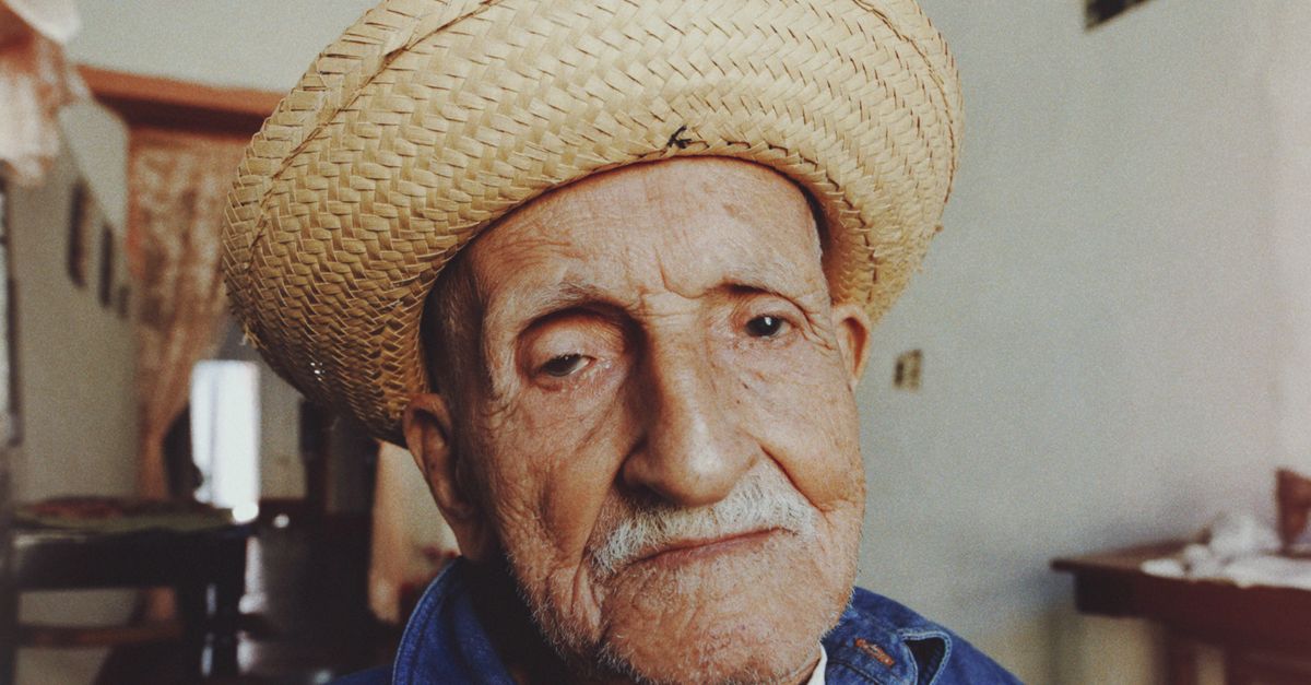 Portrait of an elderly man wearing a straw hat indoors, displaying a serene expression.