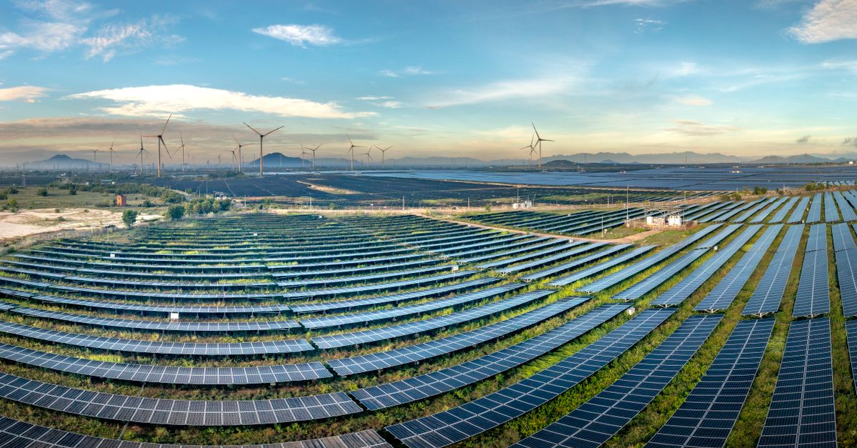 Expansive solar farm with wind turbines in the background under a clear blue sky.