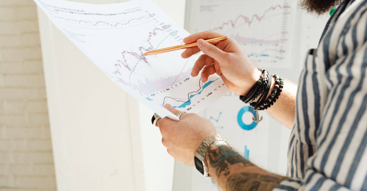 A man analyzing stock market charts with a pen, holding a paper report indoors.