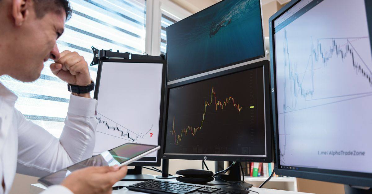 Trader in white shirt analyzing stock charts on multiple monitors during daytime in an office setting.