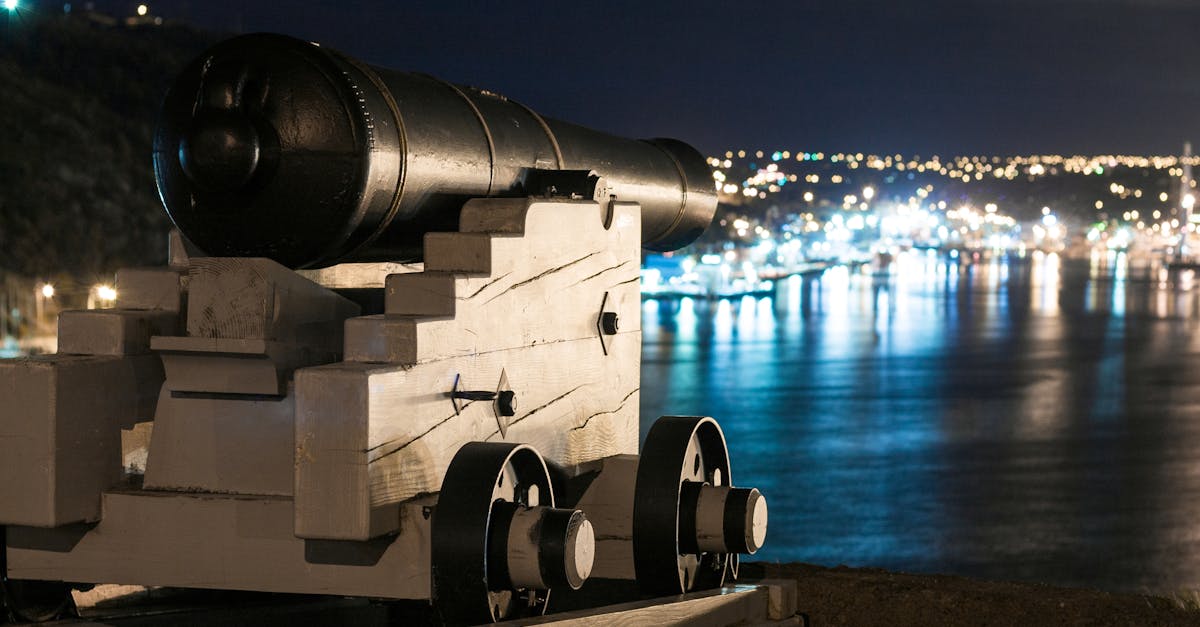 Vintage artillery cannon on wooden support on waterfront of city with glowing lights at night