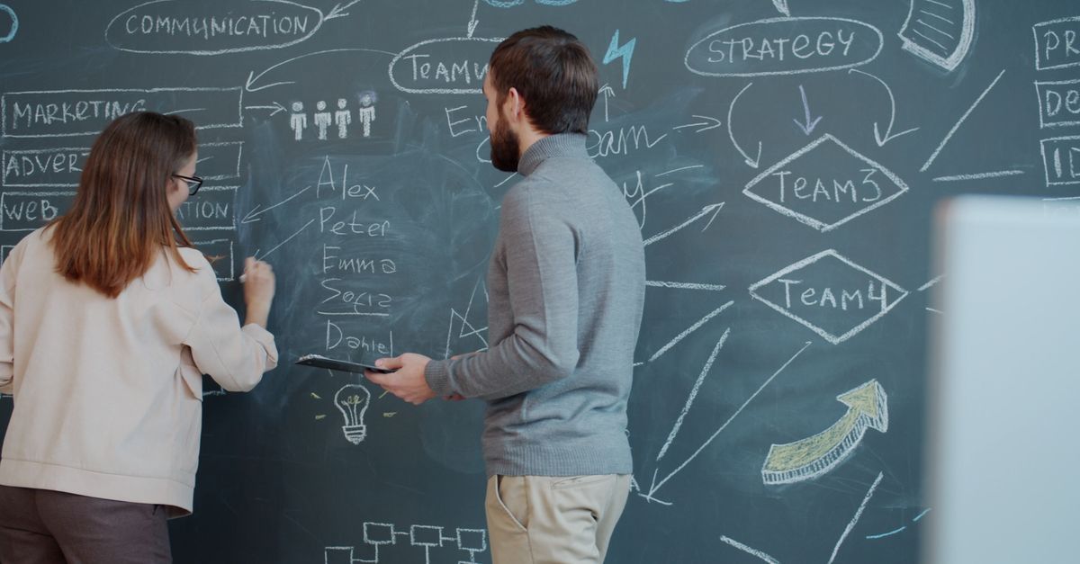 Two professionals brainstorm strategy on a chalkboard in an office setting.