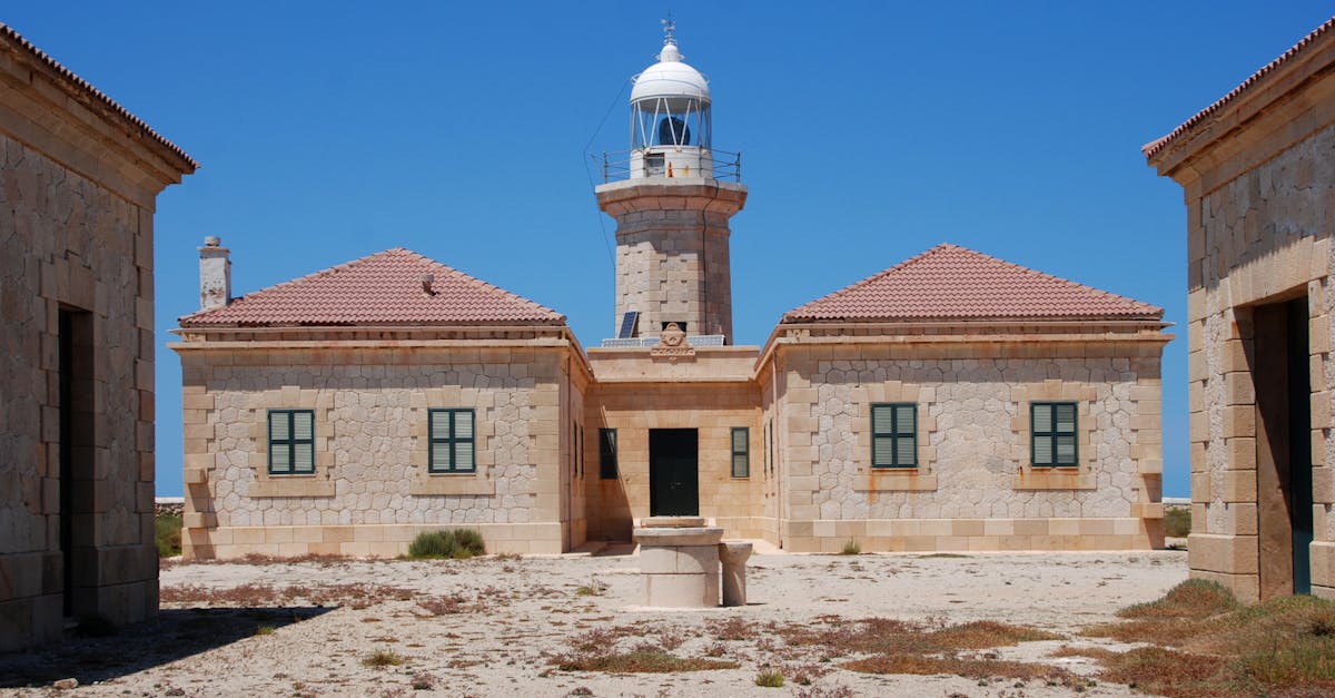 Photo of a traditional lighthouse with stone buildings in Ciutadella de Menorca, Spain, under a clear blue sky.