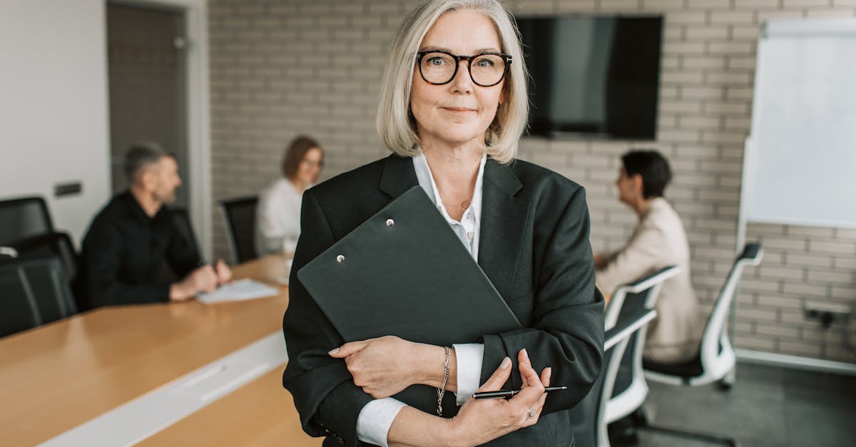 Senior businesswoman with eyeglasses holding a clipboard in a modern conference room with colleagues.