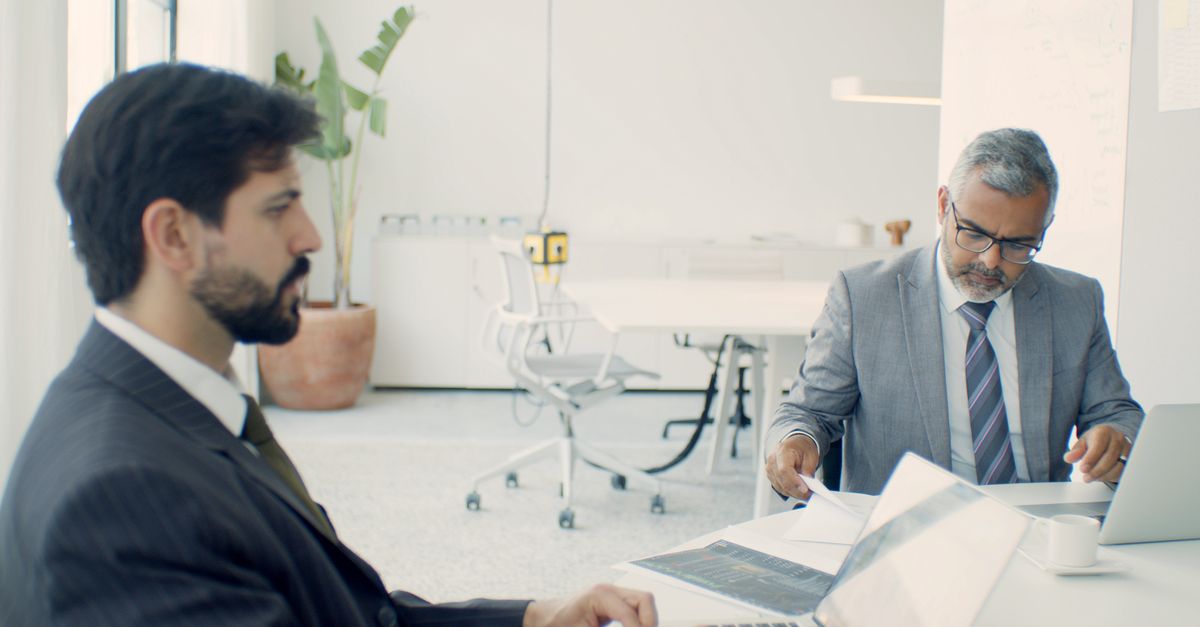 Two businessmen in formal suits working on laptops in a modern office setting.