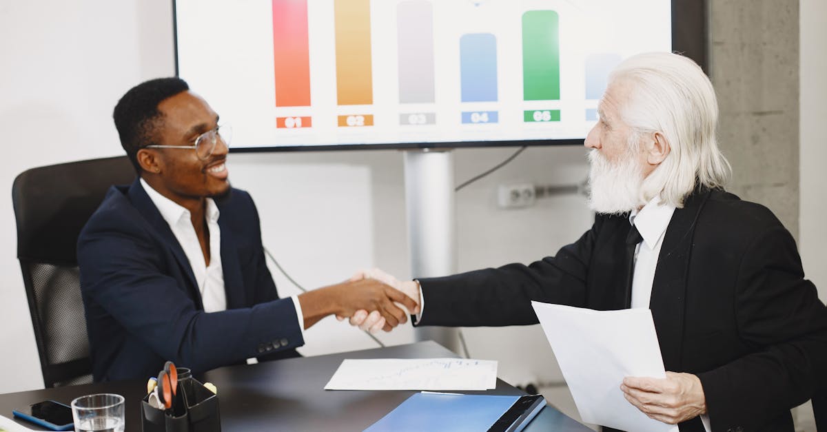 Two businessmen shaking hands in office setting with charts in background.