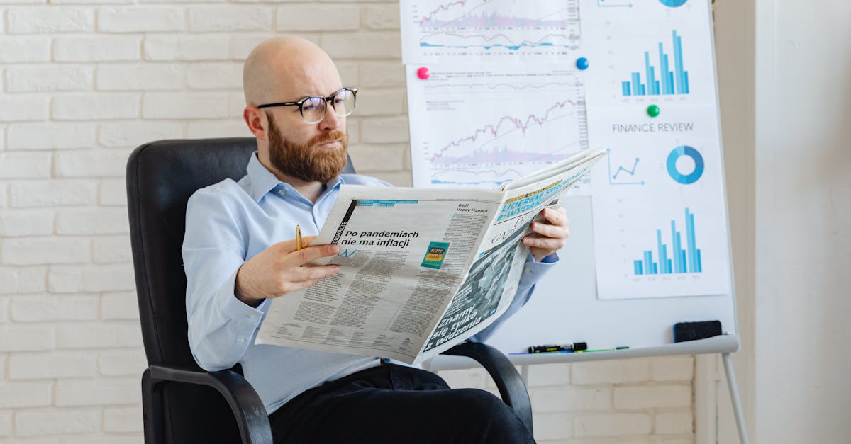 Bald man with beard reading newspaper in office with finance charts on whiteboard.
