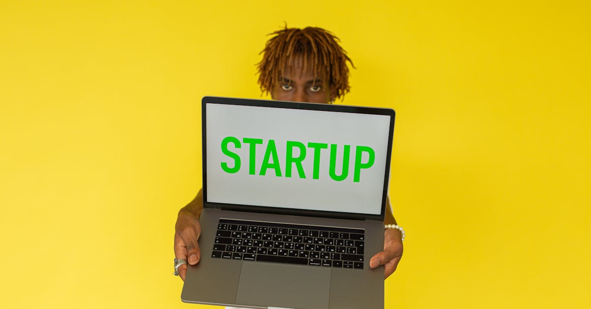 A young black man holds a laptop displaying 'Startup' against a vibrant yellow background.
